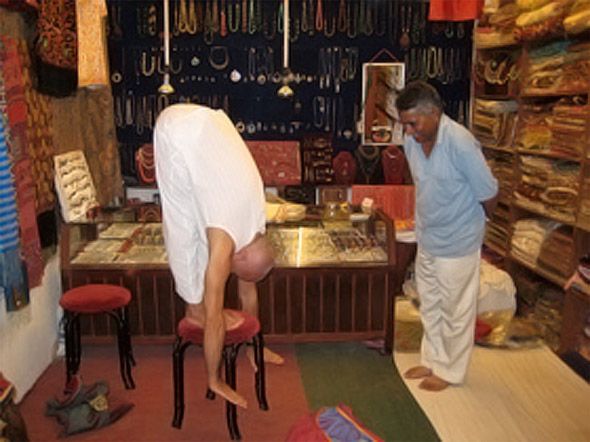 A standing forward fold practised in an Indian shop, while the shopkeeper looks on.